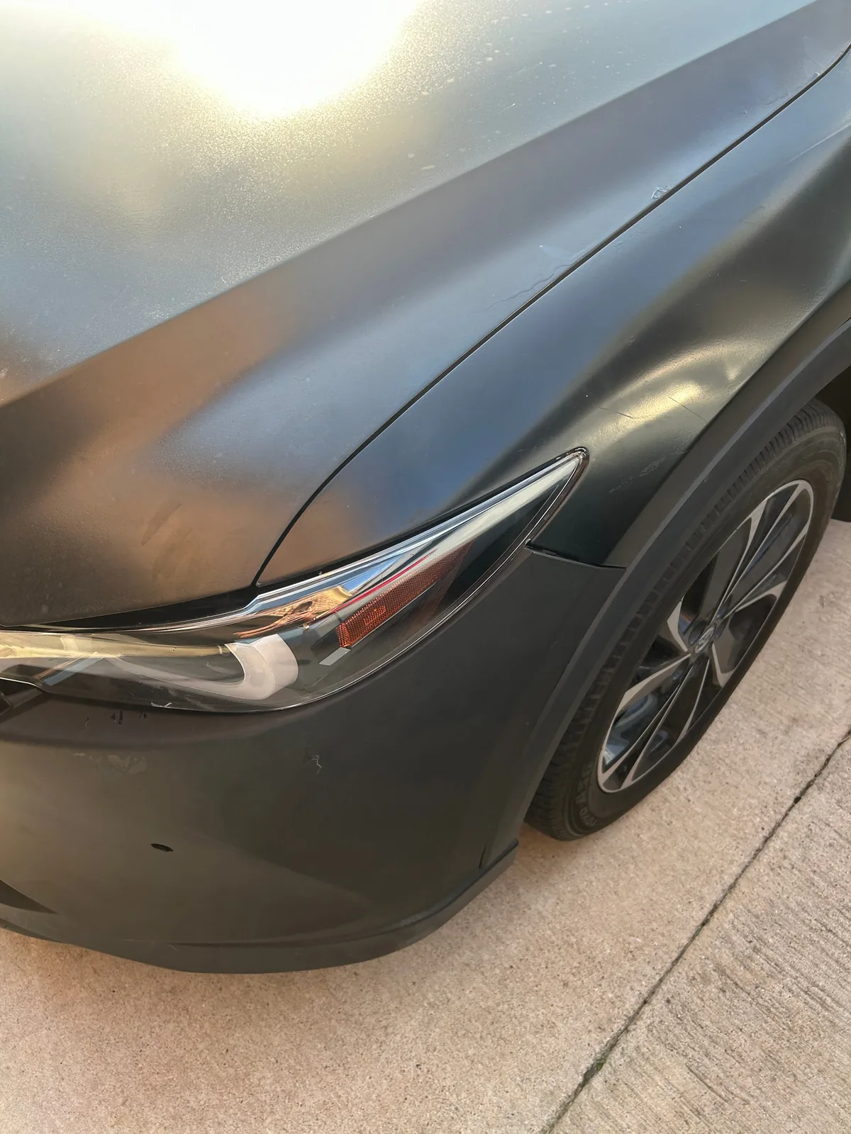 A close-up of a front corner, with the dull hood above, the fender with a few light scratches, the headlight between them and the front wheel to the right