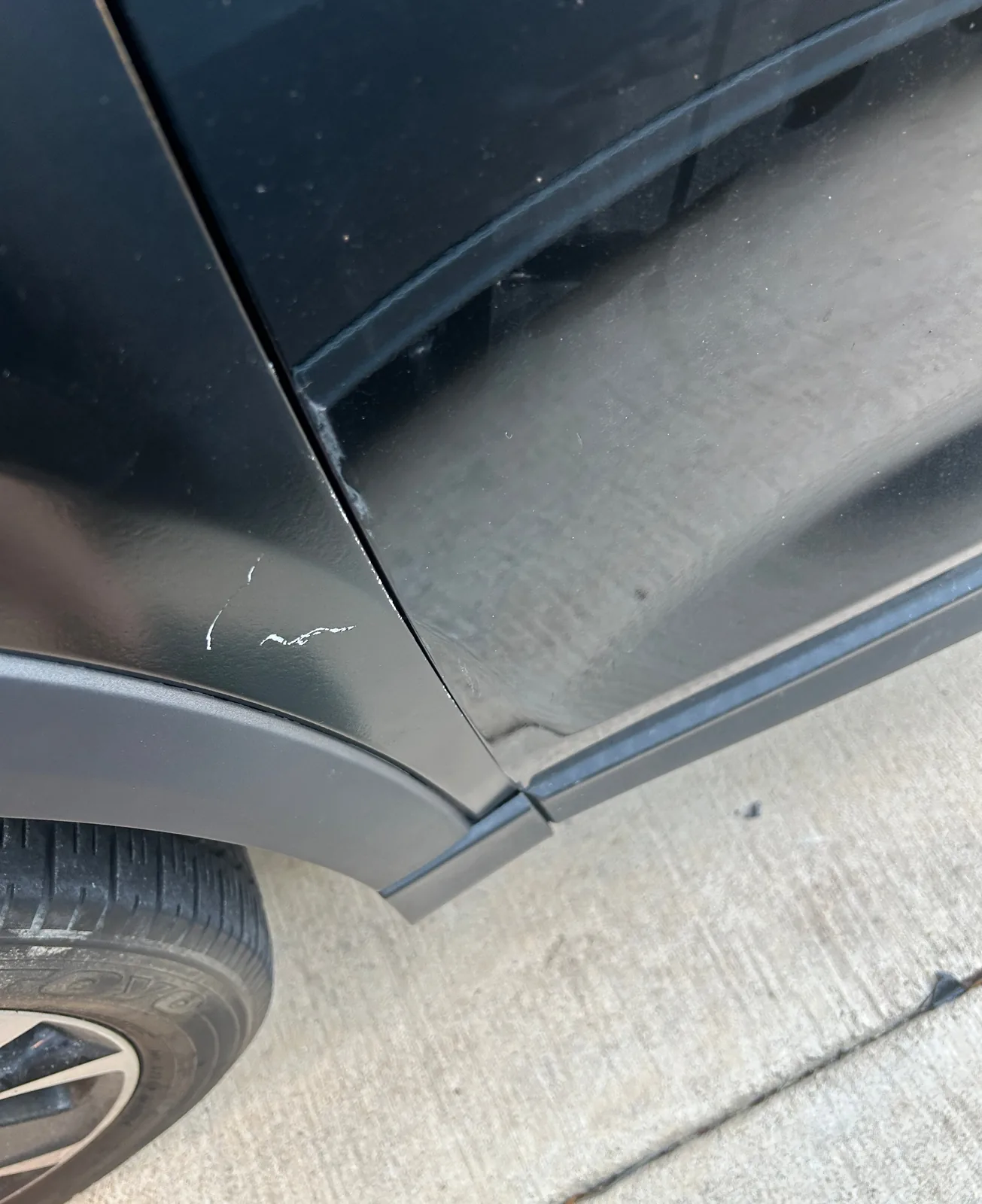 A close-up of the lower end of a panel seam, with small white scratches on the panel to its left, the black wheel arch trim and the edge of a tire at the bottom