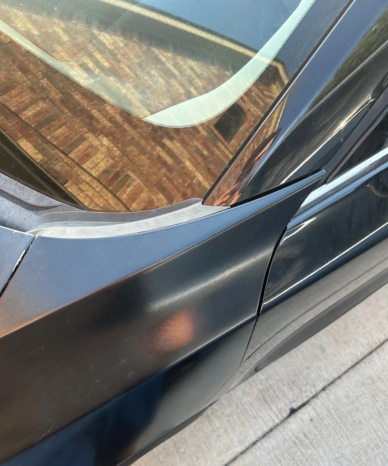 A close-up looking down at where the top of the fender meets the front door below the windshield, with a brick wall reflected in the glass