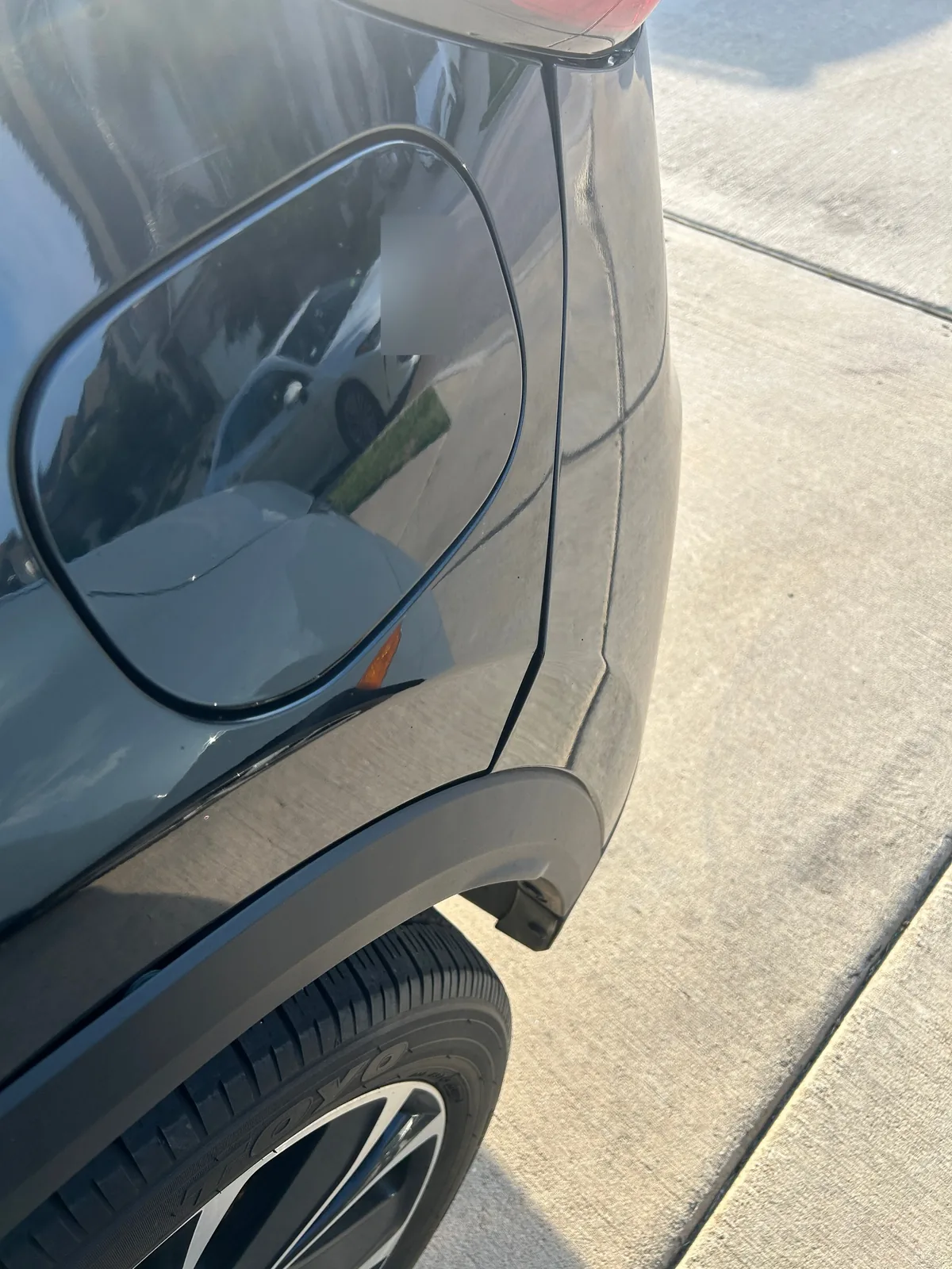 A close-up of the closed fuel door on the rear quarter panel above the rear tire, with a parked car reflected in it. The reflected car's plate is blurred