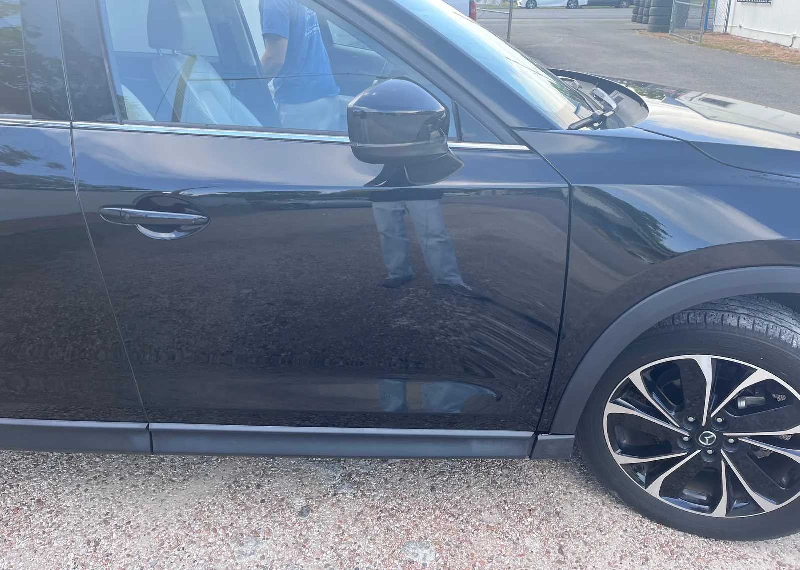 A side view of the CX-5's front right door, mirror, fender and wheel, glossy black and parked on gravel, with someone on the far side of the car seen through the window