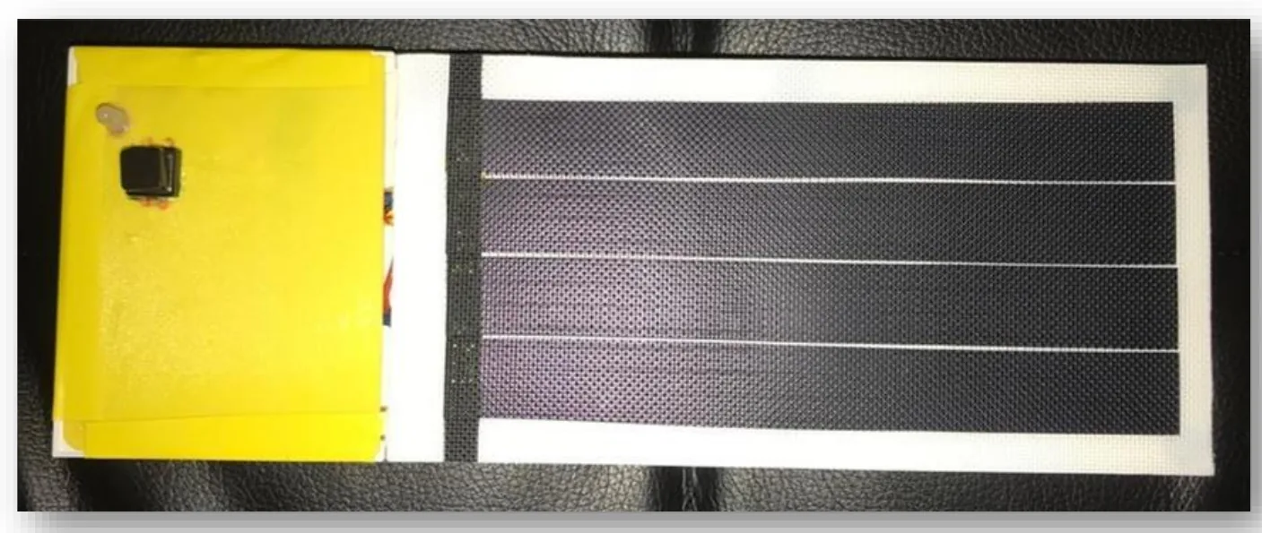A photograph of a flexible dark solar panel next to a small circuit board wrapped in yellow tape