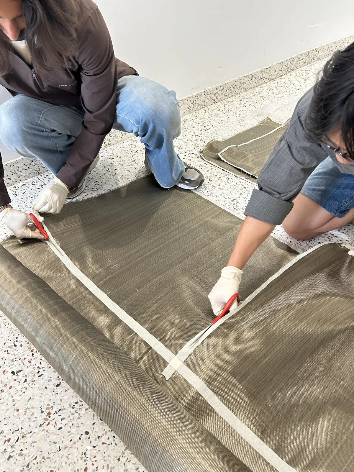 Two students in gloves kneel on a terrazzo floor, stretching a tape measure across a wide roll of olive-coloured basalt fibre fabric and cutting it with shears
