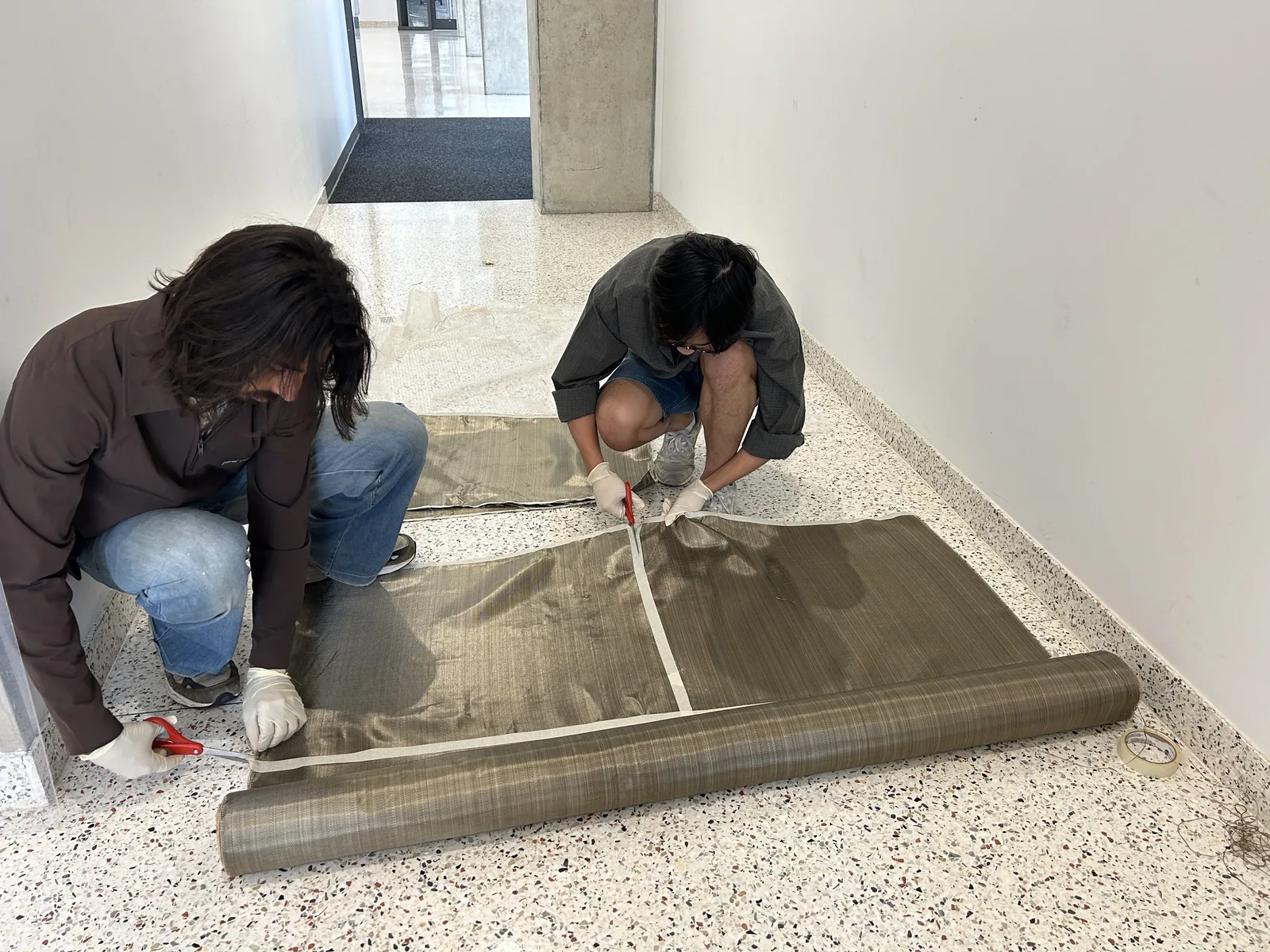 A roll of basalt fibre cloth partly unrolled along a corridor floor with two students working at one end