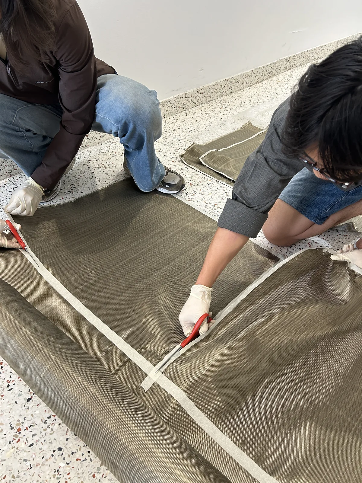 Close view of gloved hands cutting basalt fabric with red-handled shears along a measured line
