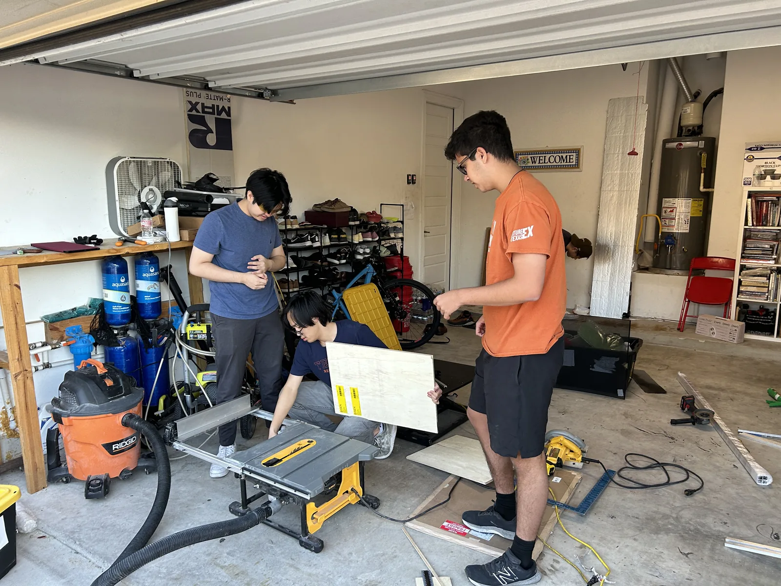 Two students cutting a large panel on a portable table saw in a domestic garage full of tools