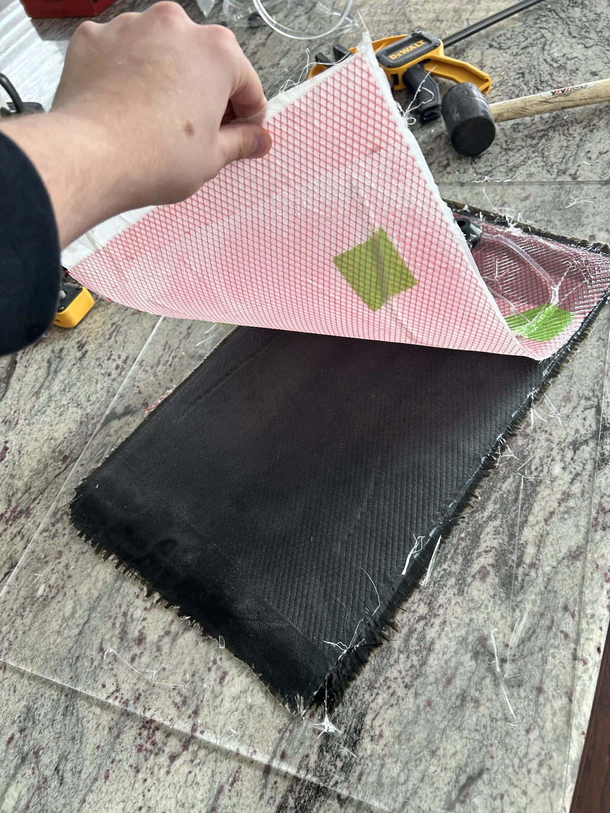 A dark composite panel on a workbench under translucent pink vacuum bagging film, with clamps and a mallet beside it