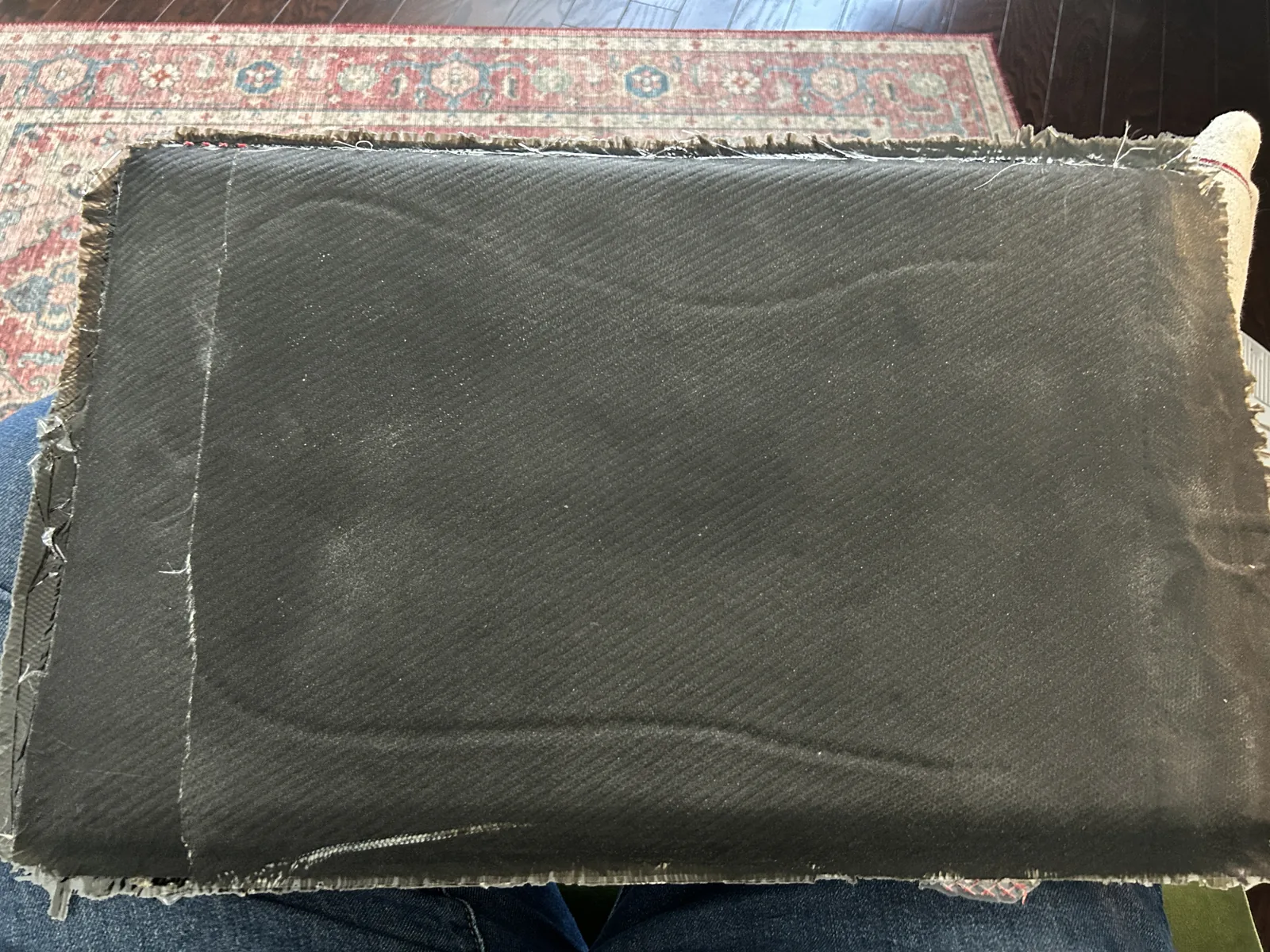 A cured flat composite test panel held up across someone's lap, showing an even grey green weave and rough trimmed edges
