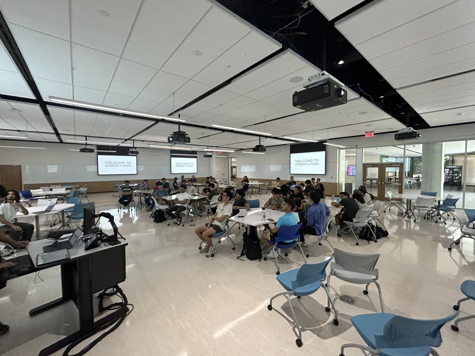 A large university classroom filled with students seated at round tables during a project meeting