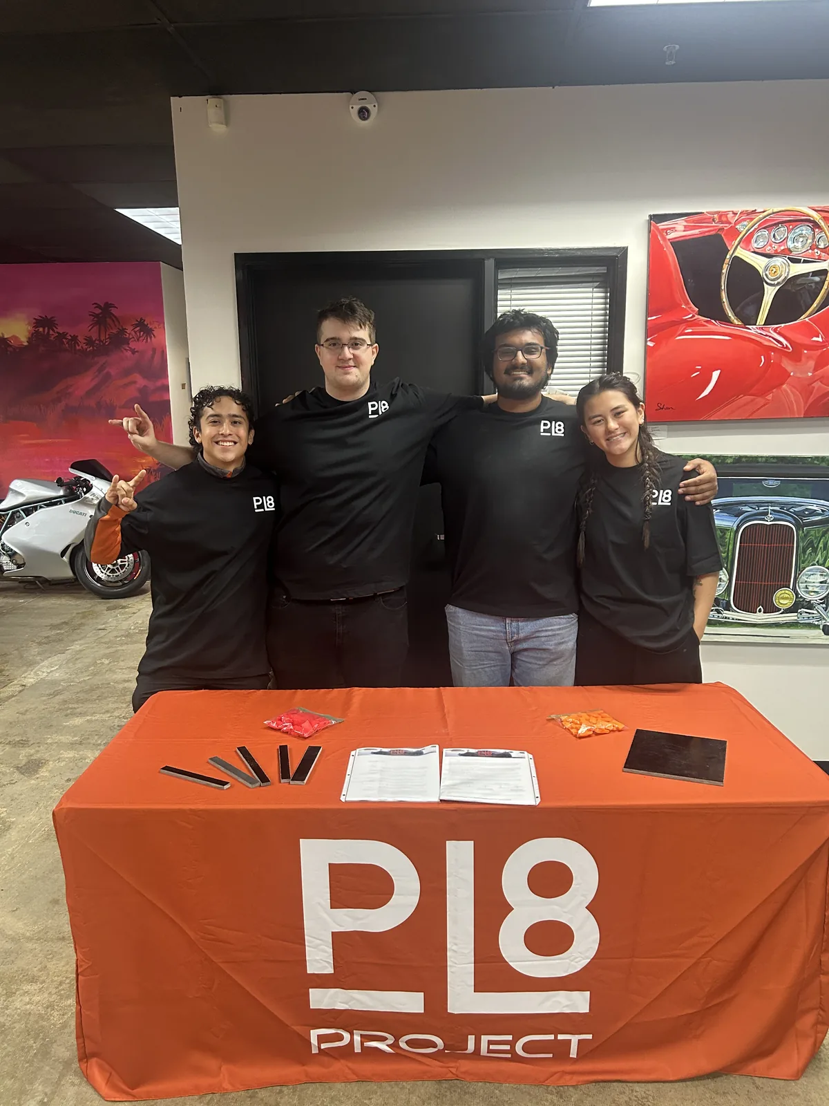 Four students in black PL8 shirts standing behind a table with an orange PL8 Project cloth