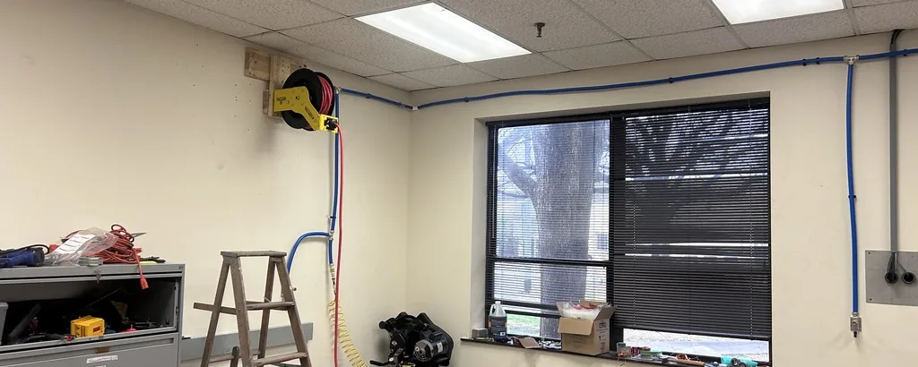 A shop room with a drop ceiling, a blue air line running along the top of the walls, a red hose reel on a yellow bracket, a window with blinds, a grey filing cabinet, a stepladder and the top of an air compressor