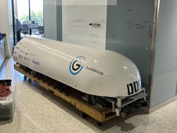 A long white capsule-shaped pod with a blue G logo and the word Guadaloop on its side, resting on a wooden pallet with casters in a hallway next to a glass-walled room