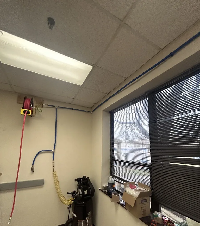 A room corner with a red hose reel high on the wall, a blue air line dropping to a fitting, a coiled yellow hose, a black upright air compressor and a window with blinds