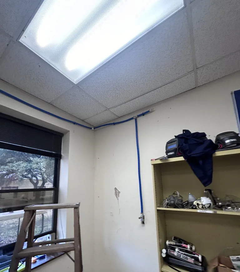 A blue air line running along the top of a wall and dropping down to a fitting, next to a window and a shelf of tools and safety gear
