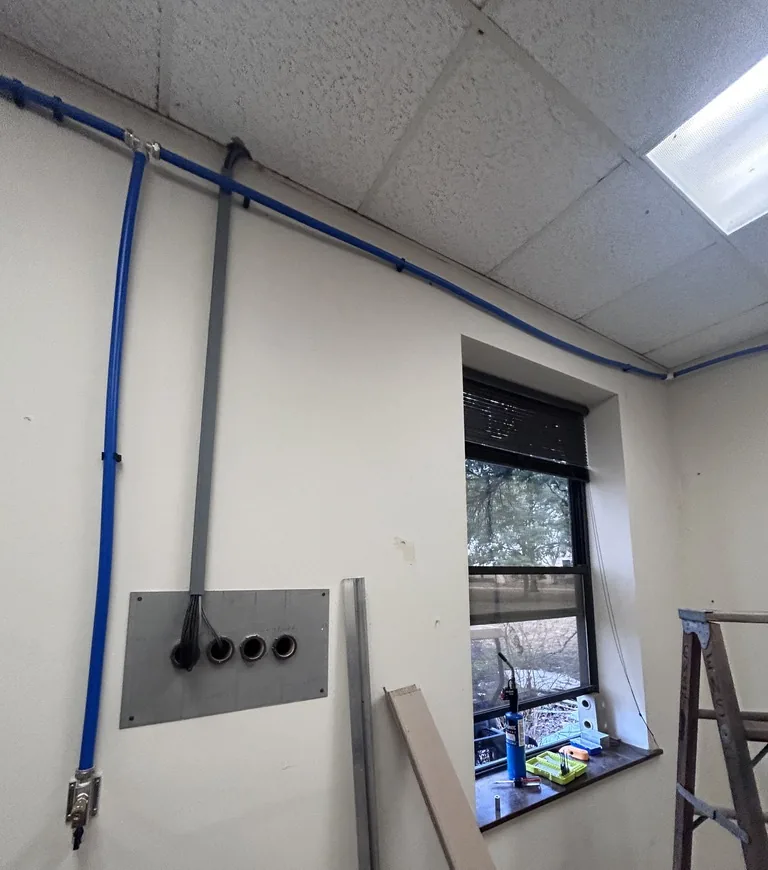 A blue air line coming down a wall beside a grey electrical conduit and a grey plate with round openings, ending in a metal fitting near the bottom of the wall