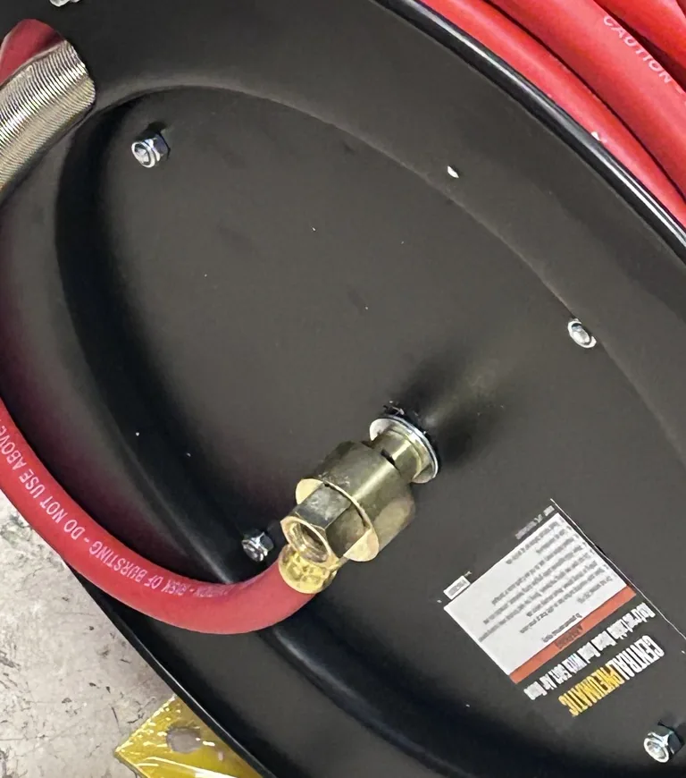The back of a black hose reel lying on a speckled floor, with a red rubber air hose wound on it and a brass fitting at the centre